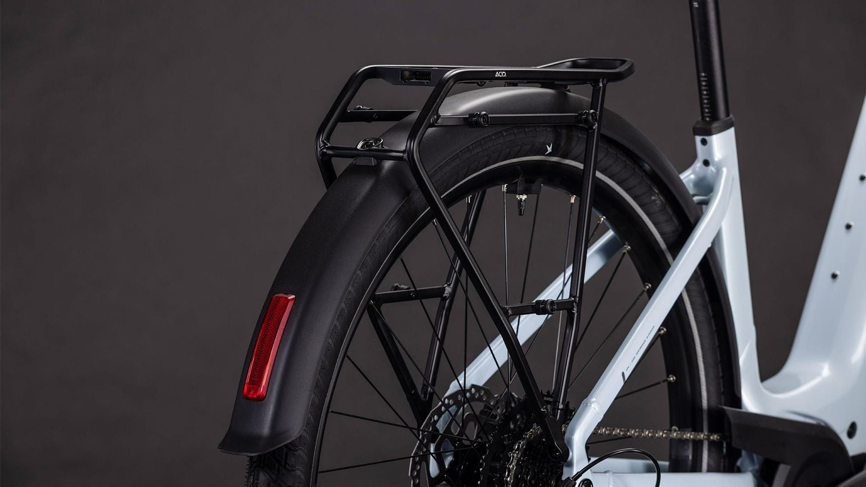Nahaufnahme des Hinterrads des CUBE Touring Hybrid ONE 600 WA mit schwarzem Schutzblech, Gangschaltung, 600-Wh-Akku und stabilem Gepäckträger vor dunklem Hintergrund. Der helle CUBE-Rahmen hebt sich elegant ab.