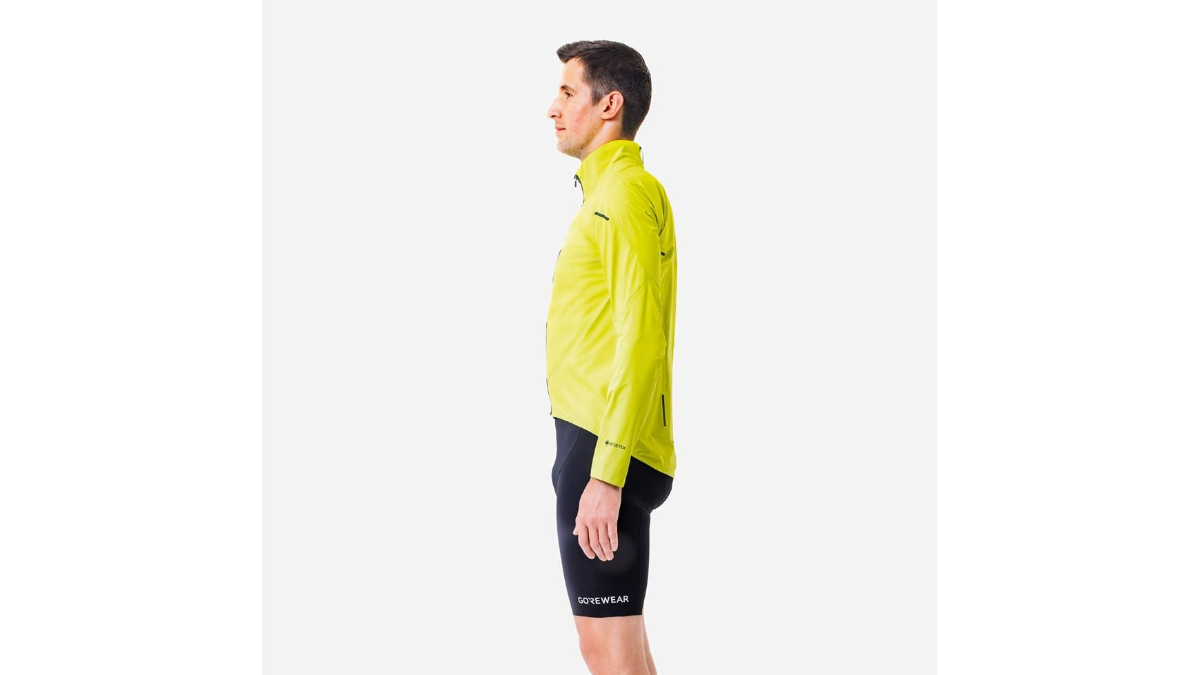 Ein Mann steht im Profil, nach links gewandt, und trägt eine leuchtend gelbe GORE SPINSHIFT GORE-TEX Jacke und schwarze Radlerhosen von GORE vor einem schlichten weißen Hintergrund.