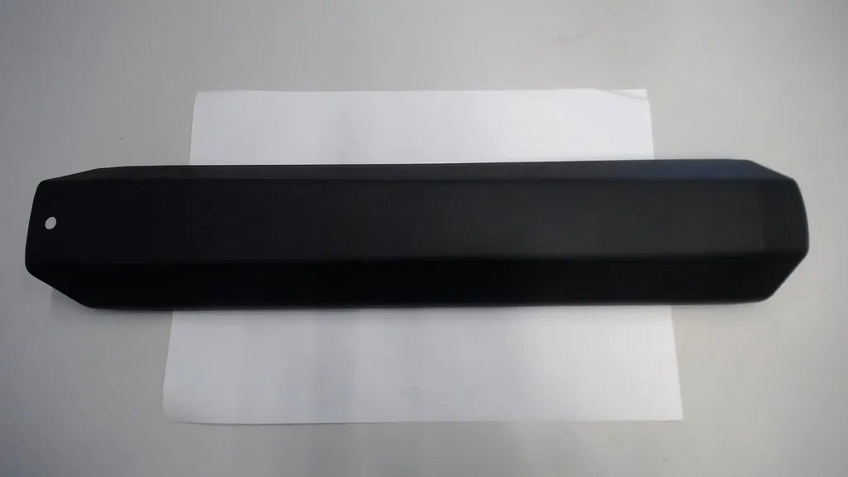 Eine schwarze, rechteckige HAIBIKE Trekking 4 Akkuabdeckung mit abgewinkelten Enden und einem kleinen kreisrunden Loch liegt mittig auf einem weißen Blatt auf einer hellgrauen Fläche und unterstreicht ihre Rolle als E-Bike-Schutzteil.